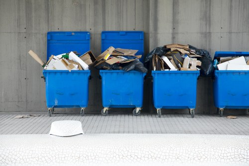 A row of five blue wheeled rubbish bins positioned on a paved area adjacent to a grey concrete wall. The bins are filled with various types of waste, including cardboard boxes, paper, and plastic packaging, some of which are overflowing and spilling out. Two of the bins are covered with black plastic rubbish bags, while others contain loose waste stacked unevenly. The environment appears to be an outdoor municipal or commercial waste disposal area, with a slight shadow cast by the bins indicating daylight. In the foreground, there is a small white object, possibly a discarded piece of paper or packaging, on the pavement. The scene is functional and typical of a rubbish collection or waste management setting in a residential or commercial vicinity, reflecting services provided by companies like House Clearance Potters Bar within the local area.