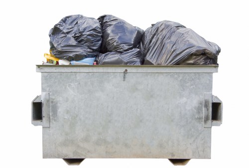 A large industrial skip bin filled with black rubbish bags, positioned outdoors on a paved surface. The bin is constructed from galvanized steel with a plain, light gray finish and features handles on each side. The black plastic bags, appearing to be filled with household or garden waste, are stacked on top of each other and vary slightly in size and shape, some leaning against the sides of the bin. The environment suggests a typical scene for rubbish collection or clearance work, possibly in a residential or commercial area within the vicinity of Potters Bar, as part of House Clearance Potters Bar’s services. The lighting indicates daylight conditions, highlighting the metallic texture of the skip and the smooth, crinkled surfaces of the plastic bags. This image exemplifies waste management practices familiar to local rubbish removal services in the area, emphasizing the process of clearing household or site waste effectively and efficiently.