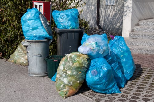 A collection of various rubbish bags and bins situated on a paved pavement outside a residential property. The refuse includes several large blue plastic bags filled with waste, and a few smaller transparent bags showing trash inside, such as discs and packaging. There are three prominent waste bins—one metallic grey, one dark grey, and one green—with the blue bags resting on or leaning against them. The items are positioned near a concrete staircase leading up to a house, with vegetation partially visible on the left side. The scene depicts an outdoor rubbish collection area, likely prepared for rubbish removal services in the Potters Bar area, with lighting suggesting daytime. House Clearance Potters Bar would organise the removal of this rubbish, helping to clear the driveway or front yard efficiently and safely, maintaining a tidy property environment.