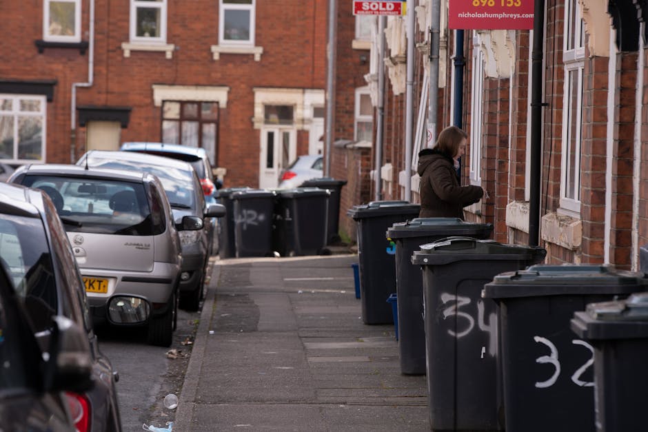 A small, old blue pickup truck with a black metal bed is parked alongside a street, loaded with a large volume of mixed rubbish including flattened cardboard boxes, plastic bags, and various discarded