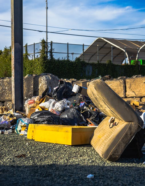 A black wheeled rubbish bin stands on a grassy patch outdoors, filled with garden waste including dried grasses, leafy green stems, and some twigs, with loose plant material spilling over the top. The